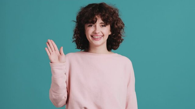 The happy curly-haired woman waving at the camera while standing in a blue studio