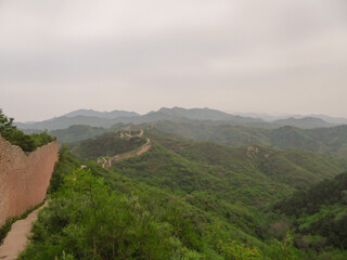 Fototapeta na wymiar A panoramic view on an unrenewed Gubeikou part of Great Wall of China. The wall is spreading on tops of mountains. Many watchtowers on the peaks. Dense forest around it. World wonder. Tradition
