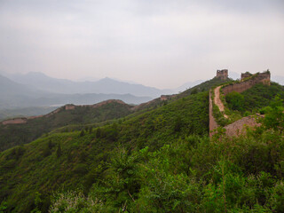 A panoramic view on an unrenewed Gubeikou part of Great Wall of China. The wall is spreading on tops of mountains. Many watchtowers on the peaks. Dense forest around it. World wonder. Tradition