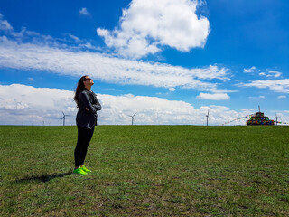 A woman standing on a vast pasture in Xilinhot in Inner Mongolia. In the back there is a heap of stones (aobao) with colorful prayer flags attached to it. Endless grassland. Blue sky with white clouds