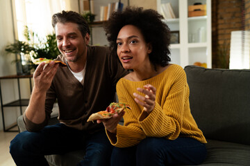 Cheerful young couple sitting on sofa at home. Happy woman and man eating pizza while watching a movie..