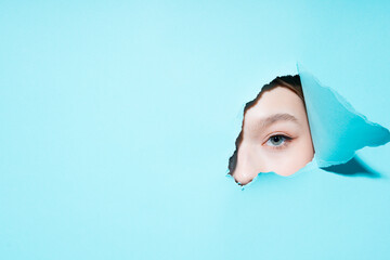 Photo of a painted eye of a girl looking at the camera through a hole on a blue background