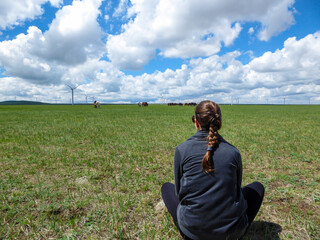 A woman with braided hair watching a heard of horses grazing under wind turbines build on a vast pasture in Xilinhot, Inner Mongolia. Natural resources. Endless grassland. Blue sky with white clouds