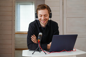 Young smiling man with headphones recording podcast using laptop at home studio, broadcasting an interview using microphone. - obrazy, fototapety, plakaty
