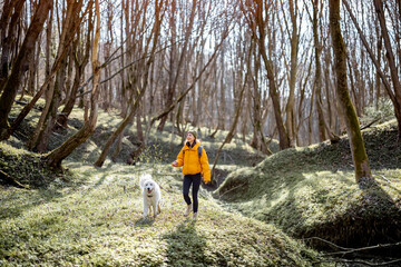 Young woman in hiking clothes and backpack spend time together with big white dog in green spring forest. Enjoys and explore of tranquil nature.