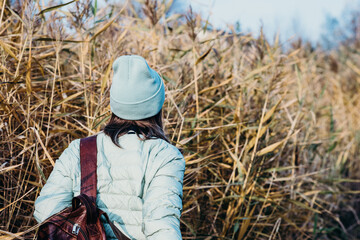 Back view of stylish girl in beige coat and hat against the background of dry reeds and blue sky. 