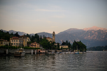 Fototapeta na wymiar Evening on lake with warm sunset colours and old italian village with church tower harbor full of yachts