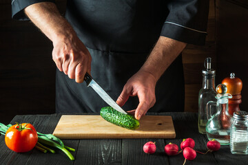 Professional chef or cook cuts green cucumber on restaurant kitchen table for salad. Vegetable diet or snack idea