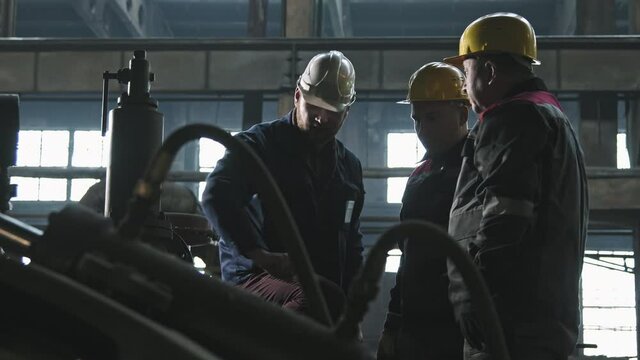 Medium shot of three male tractor plant workers in coveralls and helmets standing by machine equipment at foundry shop discussing work