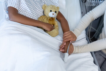 Midsection of mixed race mother and sick daughter in hospital, girl holding teddy bear