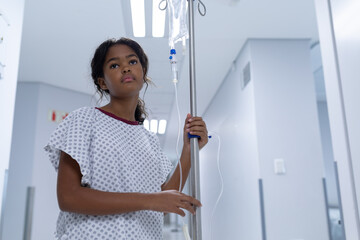 Sick mixed race girl walking in hospital corridor with iv drip bag on stand