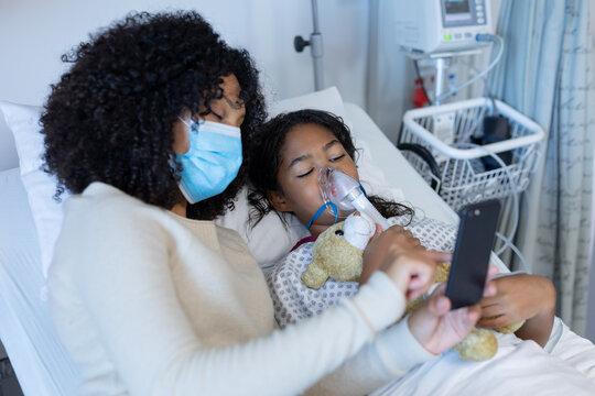 Mixed race mother in face mask with sick daughter on oxygen ventilator using smartphone in hospital