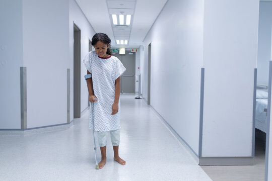 Sick Mixed Race Girl Walking Barefoot In Hospital Corridor Using A Crutch