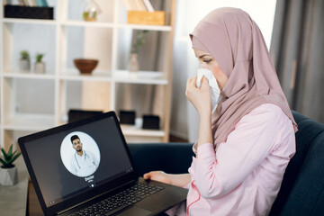 Young sick Arabian woman in hijab, suffering from cold and running nose, sits on couch at home and using laptop for video call with doctor. Female patient calling physician online.