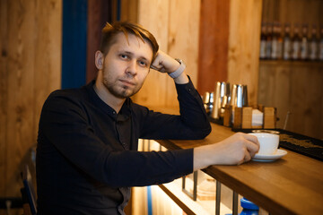 Obraz premium Close-up portrait of a young man sitting at a bar counter in a cafe with his head resting on his hand. Holds a cup of coffee and looks at the camera