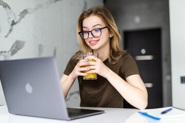 Young woman while sitting at the kitchen table, working on laptop computer, drinking juice