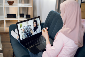 Pc monitor view over shoulder of young Muslim woman, sitting on sofa at home with thermometer . Young Arabian female doctor wear headset, gives consultation to patient about CT scan.