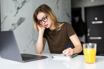 Mobile Office at home. Exhausted tired woman working using on laptop holding head in hand sitting in kitchen at home. Young girl studying or working indoors. Freelance business quarantine concept