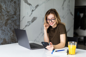 Young woman sits at the kitchen table using a laptop and use on a cell phone.