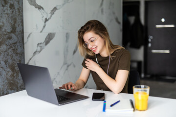 Young woman sits at the kitchen table using a laptop and use on a cell phone.