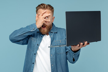 Shocked bearded blonde hipster young man looking on laptop covering eyes with hand over blue background.