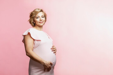 Happy smiling pregnant blonde woman stands and touches big belly. Waiting for the birth of a baby. Pink background. Space for text.