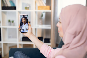 Telemedicine, online appointment concept. Side view of pretty young Muslim woman, wearing hijab, sitting on sofa at home, having video chat with female afro doctor by phone.