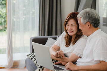 Asian senior couple video call at home. Asian Senior Chinese grandparents, using laptop video call talking with family grandchild kids while lying on sofa in living room at home concept.