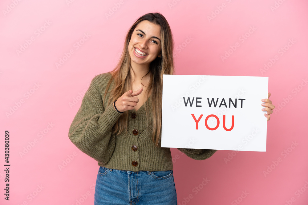 Wall mural young caucasian woman isolated on pink background holding we want you board and pointing to the fron