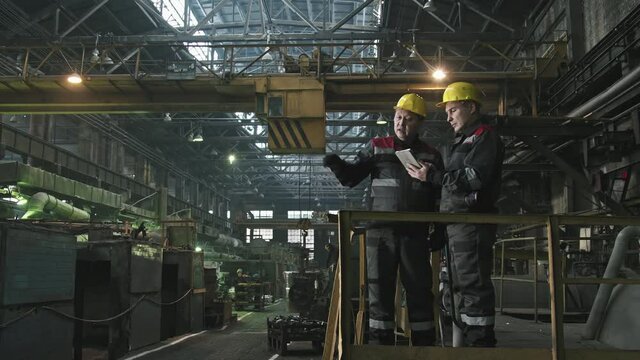 Full shot of two male tractor plant workers with digital tablet standing at foundry workshop having conversation during shift pointing at machine equipment