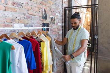 Mixed race male designer wearing tailor's meter, stacking clothes on hangers