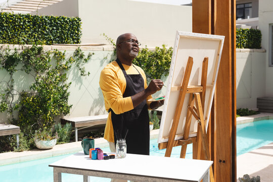 African american senior man wearing apron painting on canvas near the pool
