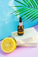 A cosmetic jar with an eyedropper stands on two granite slabs, like on a catwalk, next to half a yellow lemon, a palm leaf and the glare of the sun in the background. Cosmetics for summer