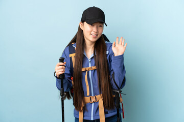 Young Chinese girl with backpack and trekking poles over isolated blue background saluting with hand with happy expression