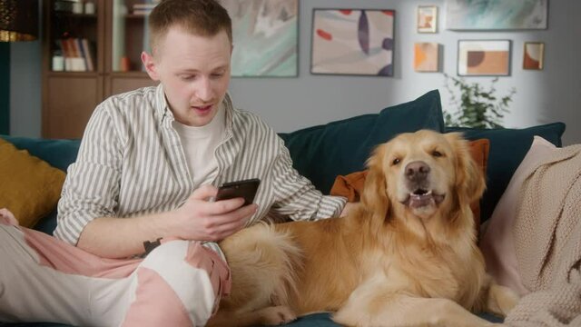 British redhead man sitting on sofa together with dog retriever making selfie photo or video call on front camera smartphone. Positive young student man conference call with pet for blog.
