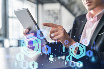 Businessman in suit using tablet device to check new candidates for international business consulting. HR, social media icons over modern panoramic office background.