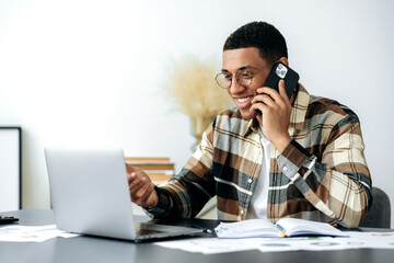 Satisfied smiling mixed race latino male freelancer or manager with glasses, have cell phone conversation, sits at work desk, uses laptop, checking e-mail, looking for information at internet