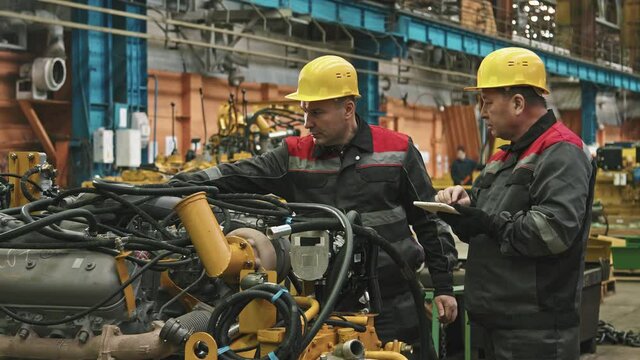 Medium shot of two male plant workers in hard hats and uniforms using digital tablet while inspecting machinery in assembly shop of tractor plant