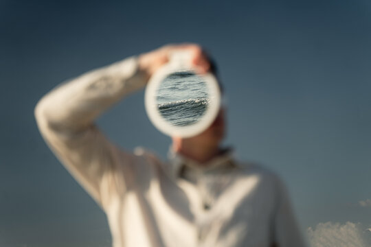 a young guy in a beige shirt holds a mirror with a reflection of the sea wave in front of his face