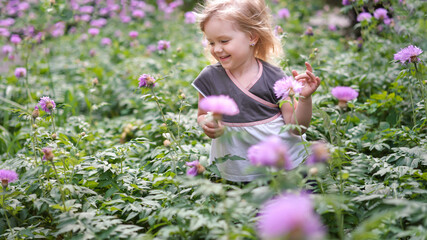 Beautiful cute little girl blonde two years old on the field in the garden in purple flowers. The child runs, jumps, has fun, grimaces in nature                     