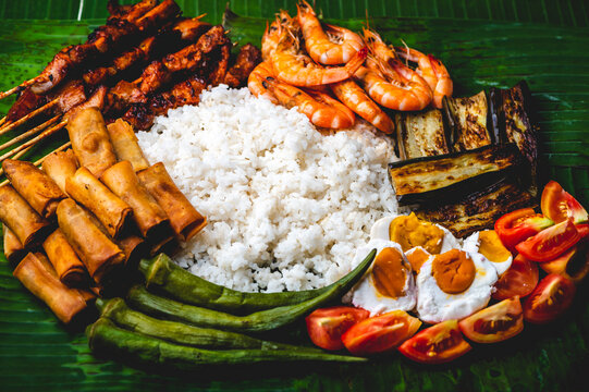 Boodle Fight- White Rice, Lumpiang Shanghai, Salted Egg, Tomato, Boiled Okra, Fried Eggplant, Shrimps, and Pork barbecue (flat lay close up)