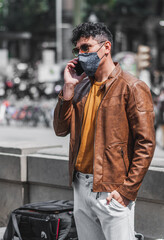 Good looking young adult man wearing casual spring clothes outdoor,  talking and texting on phone in padendemic day taking measure with face mask.