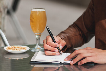 Young man in fashion spring clothes. On his weekend, relaxed in city having a beer and drawing  under trees, on a table terrace in the bar