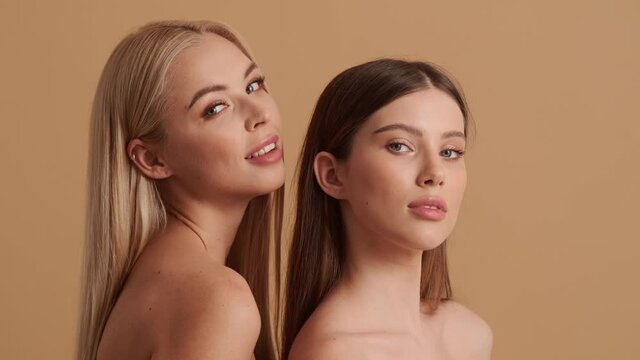 Charming sexy young half-naked women are posing to the camera standing isolated over a beige background in the studio