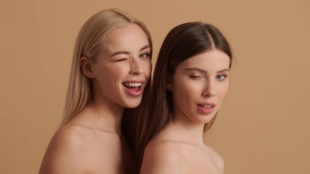 Beautiful young half-naked women are winking while looking to the camera standing isolated over a beige background in the studio