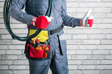 Electrician with tools wearing uniform holding rolled wires and showing thumb up on brick wall background.
