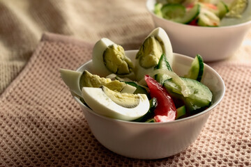 Salad with chicken eggs and fresh vegetables in white plates. Cucumbers and tomatoes in slices, on the table cloth texture napkins. Vintage wall at the back