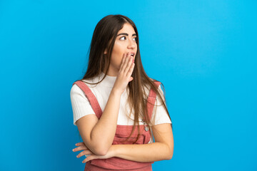 Young caucasian woman isolated on blue background whispering something with surprise gesture while looking to the side