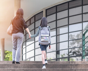 Educational back to school, parent's day, bring kid to work concept with elementary student girl child carrying backpack holding mother's hand walking up admission office building going to class