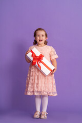 little cute girl with gift box with red ribbon on purple background. birthday of small child. a happy child holds a gift in his hands. a birthday present. concept of celebrating, giving and receiving